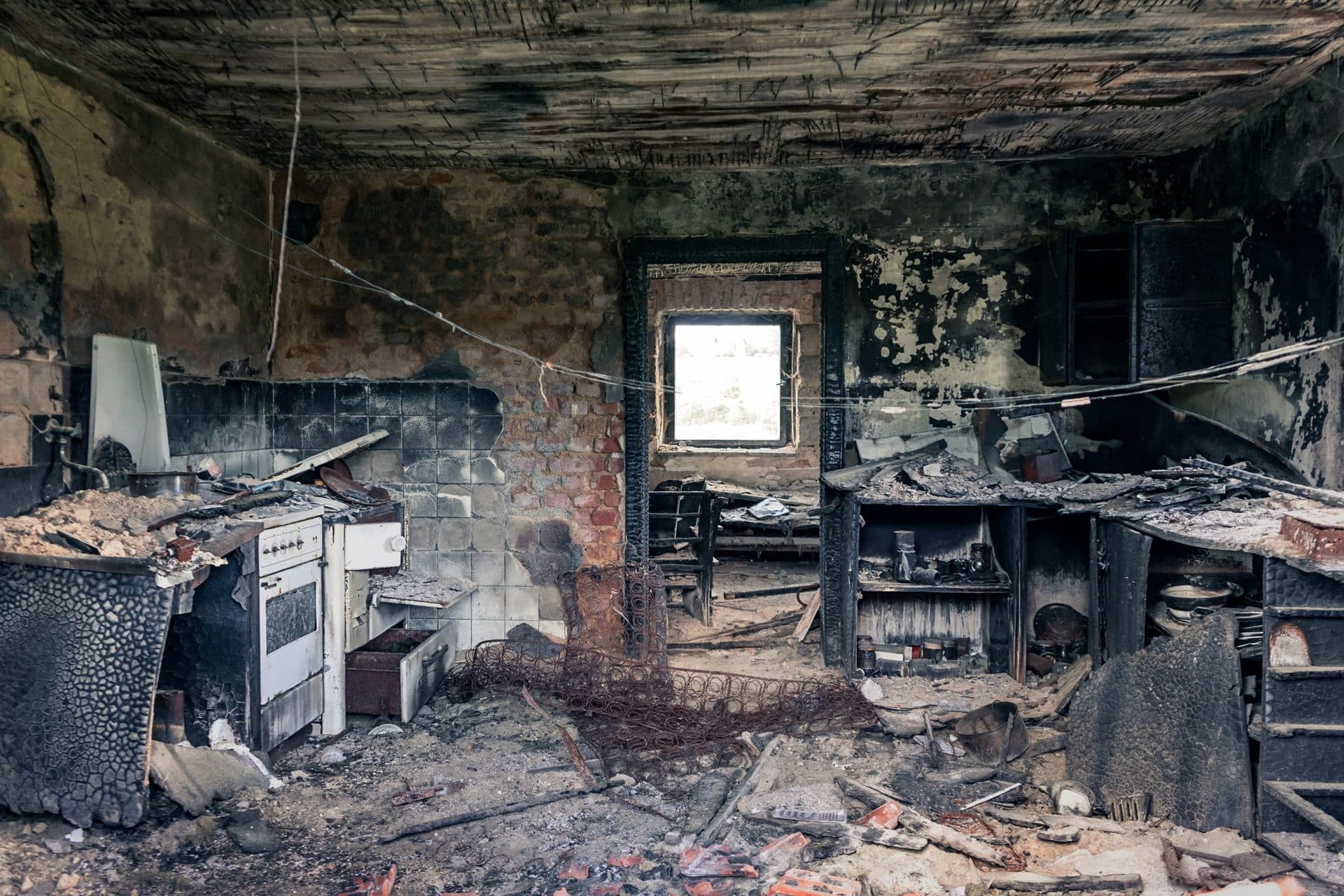 A kitchen gutted by fire, with soot blackening across the whole ceiling, burnt-out cabinets and a scorched cooker, and charred debris across the floor