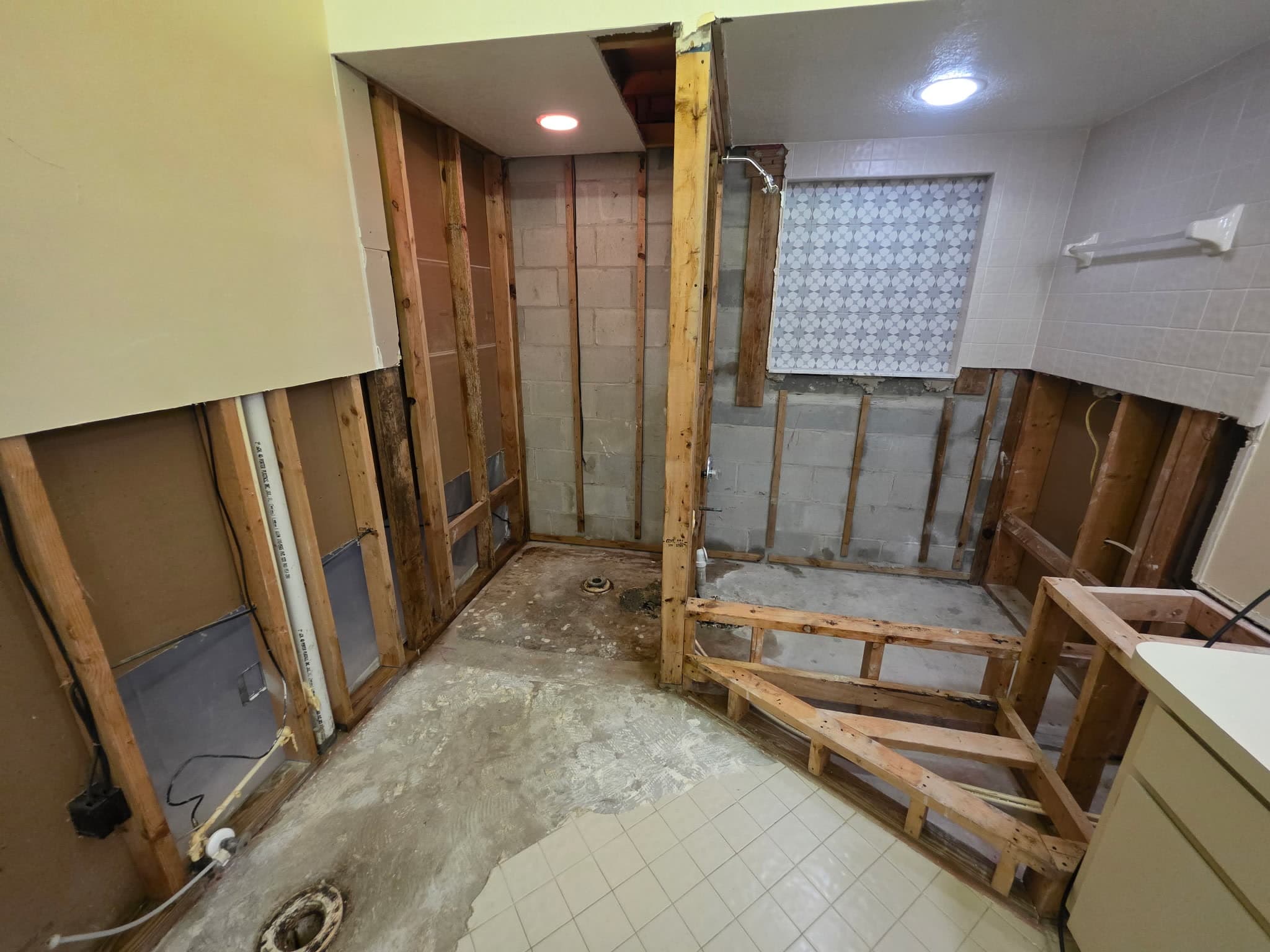 A bathroom gutted back to studs and concrete block, with the tub deck framing exposed, the closet flange open in the slab and the old vanity still standing