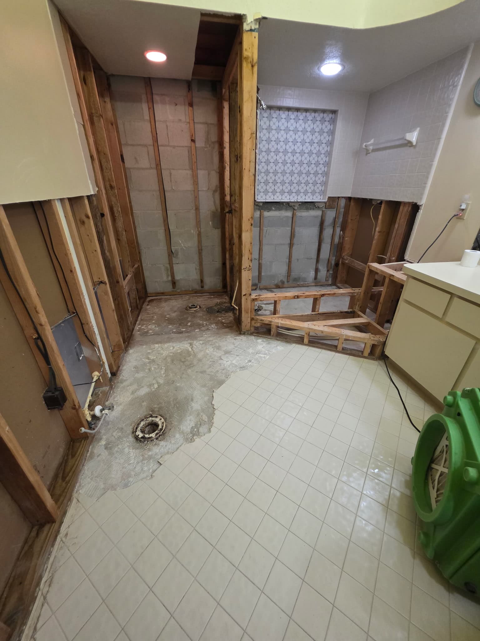 An air mover running on the tile floor of a bathroom stripped back to studs and block, drying the room before anything is rebuilt