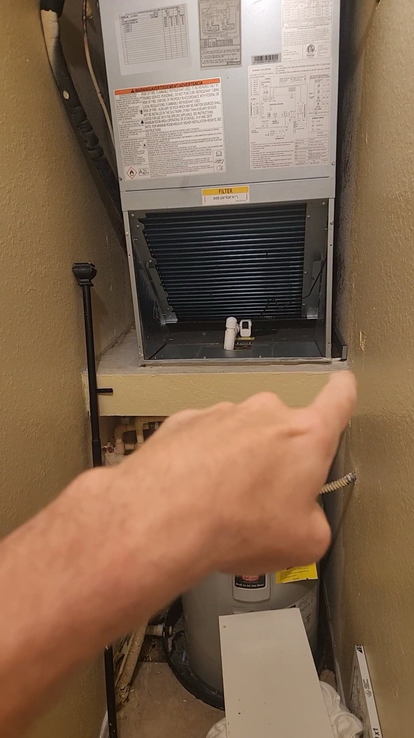A hand pointing into an open air handler closet, at the coil compartment above the water heater, during an inspection